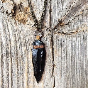 Puede incluir: Un colgante de punta de flecha de obsidiana negra en una cadena de collar de bronce. La punta de flecha est&aacute; envuelta con cord&oacute;n marr&oacute;n y cuelga de un anillo de bronce.