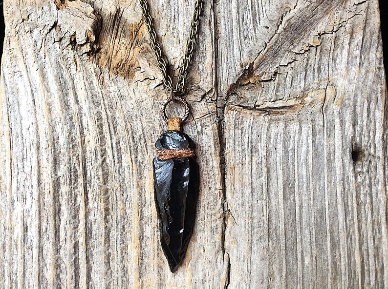 Puede incluir: Un colgante de punta de flecha de obsidiana negra en una cadena de collar de bronce. La punta de flecha est&aacute; envuelta con cord&oacute;n marr&oacute;n y cuelga de un anillo de bronce.