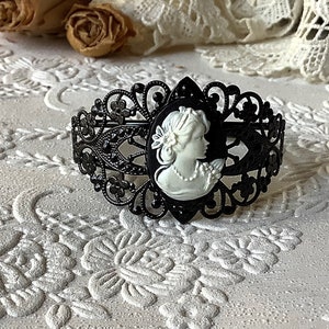May include: A black filigree bracelet with a white cameo of a woman's profile. The bracelet is on a white background with a floral pattern.
