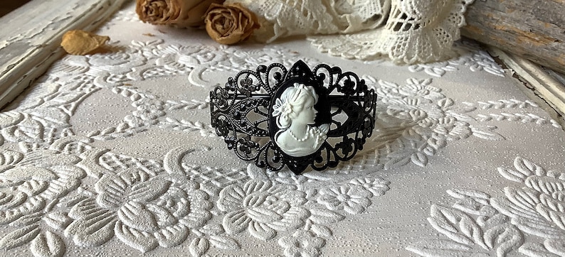 May include: A black filigree bracelet with a white cameo of a woman's profile. The bracelet is on a white background with a floral pattern.