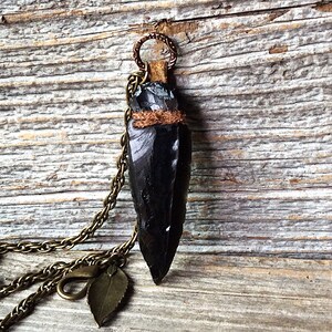 Puede incluir: Un colgante de punta de flecha de obsidiana negra en una cadena de lat&oacute;n con un dije de hoja. La punta de flecha est&aacute; envuelta en cuero marr&oacute;n y tiene un acento de alambre de cobre.