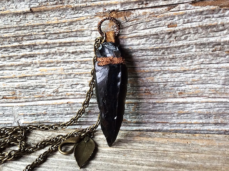 Puede incluir: Un colgante de punta de flecha de obsidiana negra en una cadena de lat&oacute;n con un dije de hoja. La punta de flecha est&aacute; envuelta en cuero marr&oacute;n y tiene un acento de alambre de cobre.