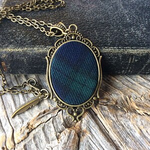 May include: A bronze chain necklace with a green and blue plaid fabric pendant in an ornate oval frame. The pendant is attached to a small bronze feather charm.