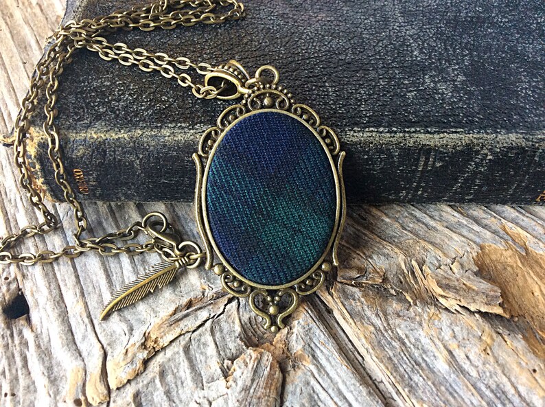 May include: A bronze chain necklace with a green and blue plaid fabric pendant in an ornate oval frame. The pendant is attached to a small bronze feather charm.