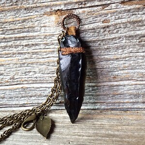 Puede incluir: Un colgante de punta de flecha de obsidiana negra en una cadena de lat&oacute;n con un dije de hoja. El colgante est&aacute; envuelto con cord&oacute;n de cuero marr&oacute;n.