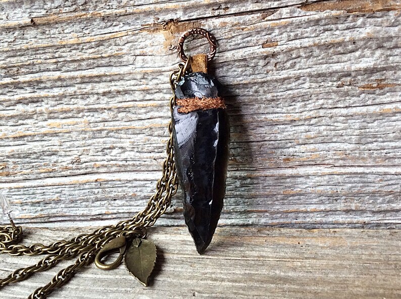 Puede incluir: Un colgante de punta de flecha de obsidiana negra en una cadena de lat&oacute;n con un dije de hoja. El colgante est&aacute; envuelto con cord&oacute;n de cuero marr&oacute;n.