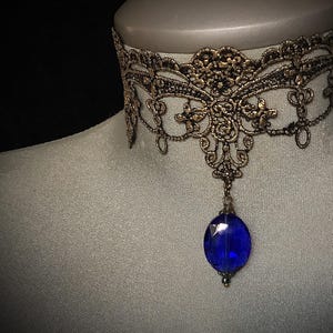 Pode incluir: Um colar de renda dourado com um pingente de pedra preciosa azul.