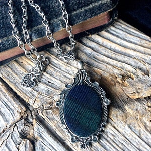 May include: A silver chain necklace with a heart charm and a pendant featuring a dark green and black patterned fabric inside a silver ornate frame.