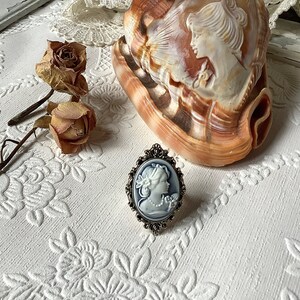 May include: A silver ring with a white cameo featuring a woman's profile. The cameo is set in an oval frame with ornate details. The ring is on a white surface with a floral pattern.