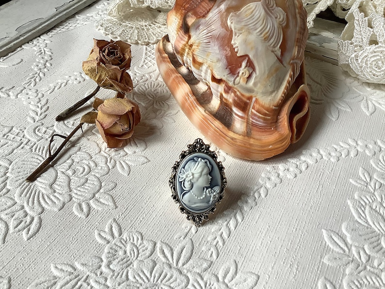 May include: A silver ring with a white cameo featuring a woman's profile. The cameo is set in an oval frame with ornate details. The ring is on a white surface with a floral pattern.