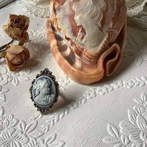 May include: A silver ring with a blue cameo featuring a woman's profile. The ring is on a white surface with a floral pattern.