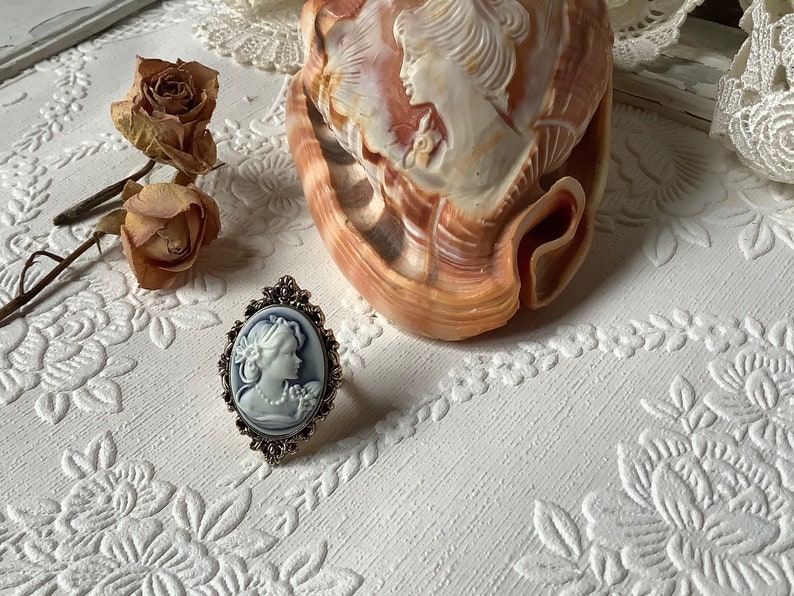 May include: A silver ring with a blue cameo featuring a woman's profile. The ring is on a white surface with a floral pattern.