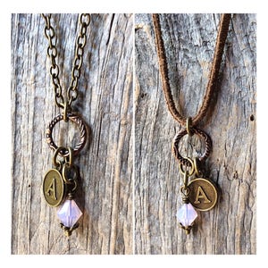 May include: Two necklaces with a charm featuring the letter A, a small crystal, and a twisted ring. One necklace has a brown leather cord and the other has a silver chain.