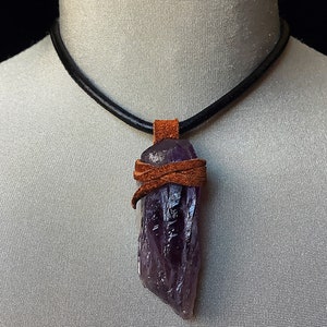 Puede incluir: Un colgante de cristal de amatista púrpura envuelto en un cordón de cuero marrón en un collar de cuero negro.