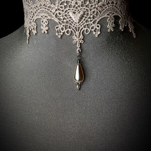 May include: A delicate lace choker necklace with a pearlescent teardrop pendant. The choker is a light beige color and the pendant is a soft white.