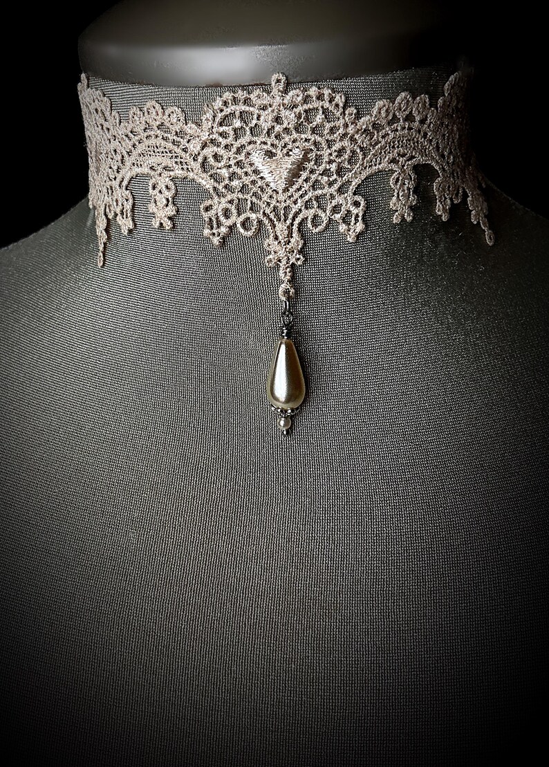 May include: A delicate lace choker necklace with a pearlescent teardrop pendant. The choker is a light beige color and the pendant is a soft white.
