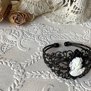 May include: Black filigree bracelet with a white cameo portrait. The bracelet is on a white surface with a floral pattern.