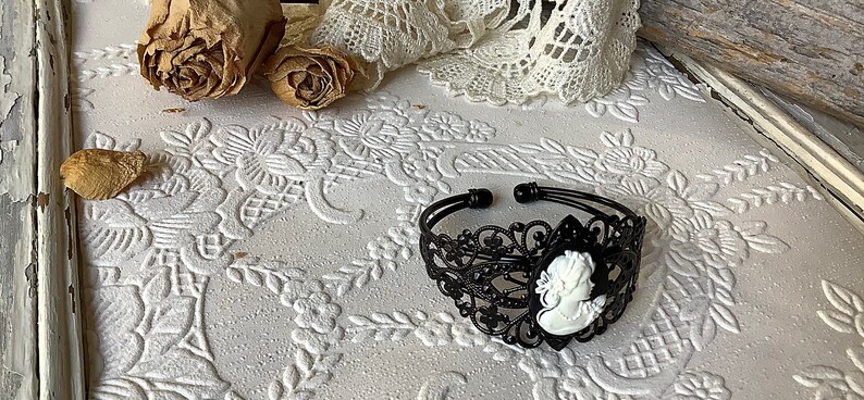 May include: Black filigree bracelet with a white cameo portrait. The bracelet is on a white surface with a floral pattern.