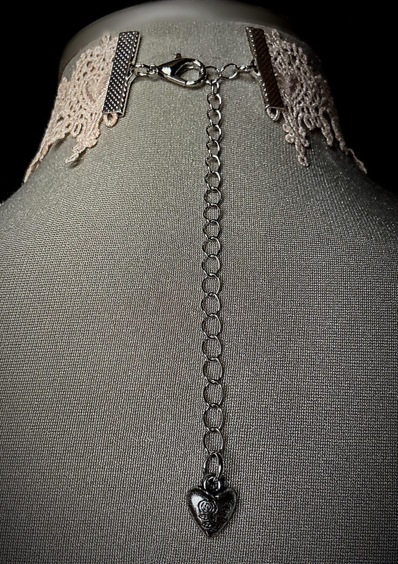 May include: A delicate white lace choker with a silver chain and a heart-shaped pendant. The pendant has a small rose design.