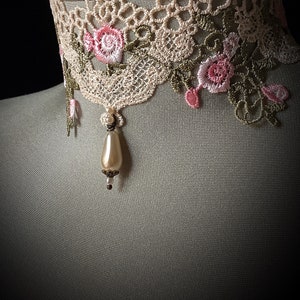 Puede incluir: Un delicado collar de encaje marfil con bordados florales rosas y verdes y un colgante de perla.