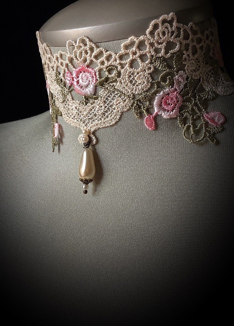 Puede incluir: Un delicado collar de encaje marfil con bordados florales rosas y verdes y un colgante de perla.