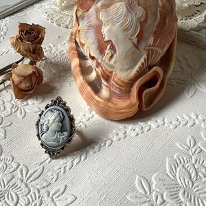 May include: A silver ring with a cameo of a woman's face in a white oval setting. The ring is on a white surface with a seashell and dried roses.