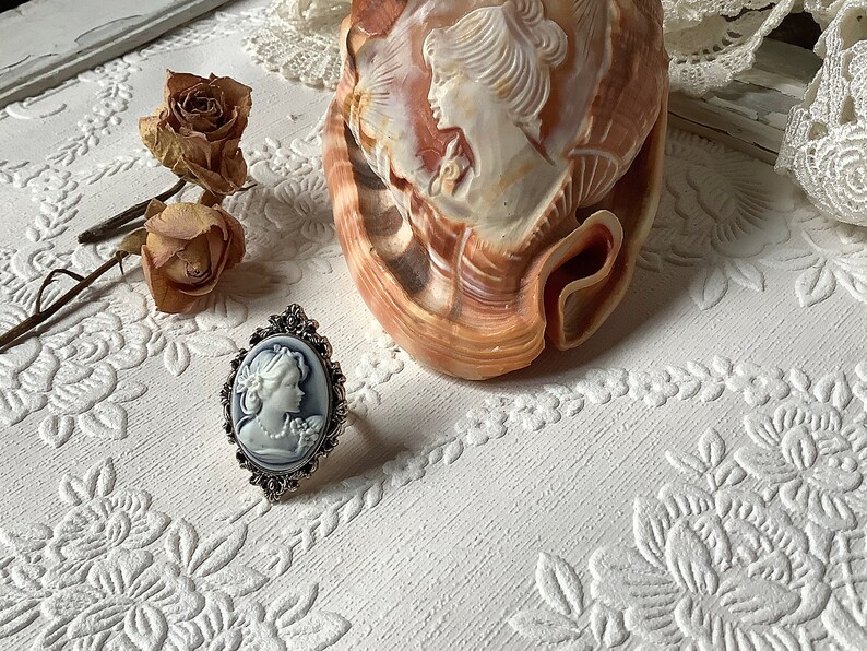 May include: A silver ring with a cameo of a woman's face in a white oval setting. The ring is on a white surface with a seashell and dried roses.