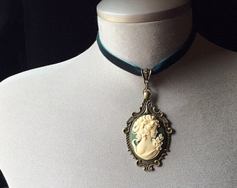 Może przedstawiać: Czarny aksamitny choker z pozłacanym wisiorem, na którym znajduje się kamea z profilem kobiety. Kamea jest osadzona w ozdobnej ramie z zielonym tłem.