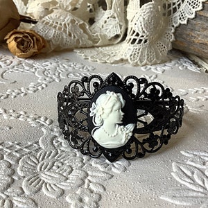 May include: Black metal bracelet with a white cameo featuring a woman's profile. The bracelet has a delicate, intricate design.