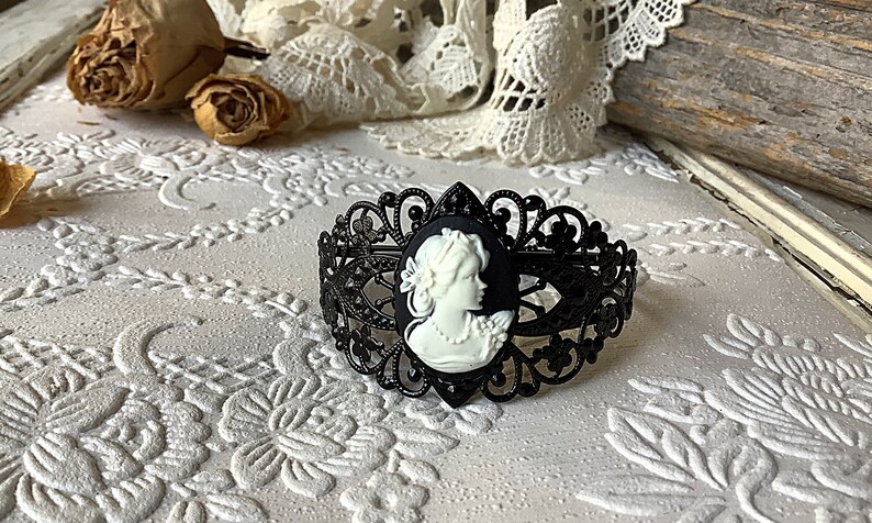 May include: Black metal bracelet with a white cameo featuring a woman's profile. The bracelet has a delicate, intricate design.