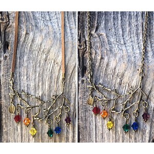 May include: A bronze-toned necklace featuring a branch design with colorful, faceted crystal pendants in a rainbow spectrum. The necklace has a suede cord and a chain option. The background is a weathered wood surface.