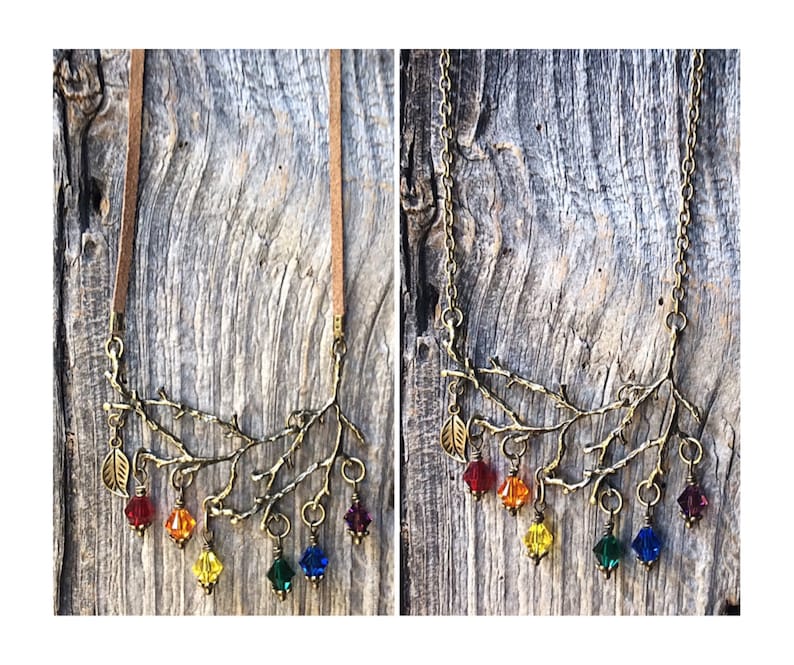 May include: A bronze-toned necklace featuring a branch design with colorful, faceted crystal pendants in a rainbow spectrum. The necklace has a suede cord and a chain option. The background is a weathered wood surface.