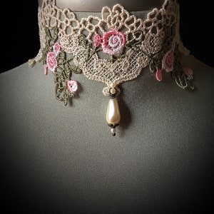 Puede incluir: Un delicado collar de encaje beige con bordado floral rosa y un colgante de perla.