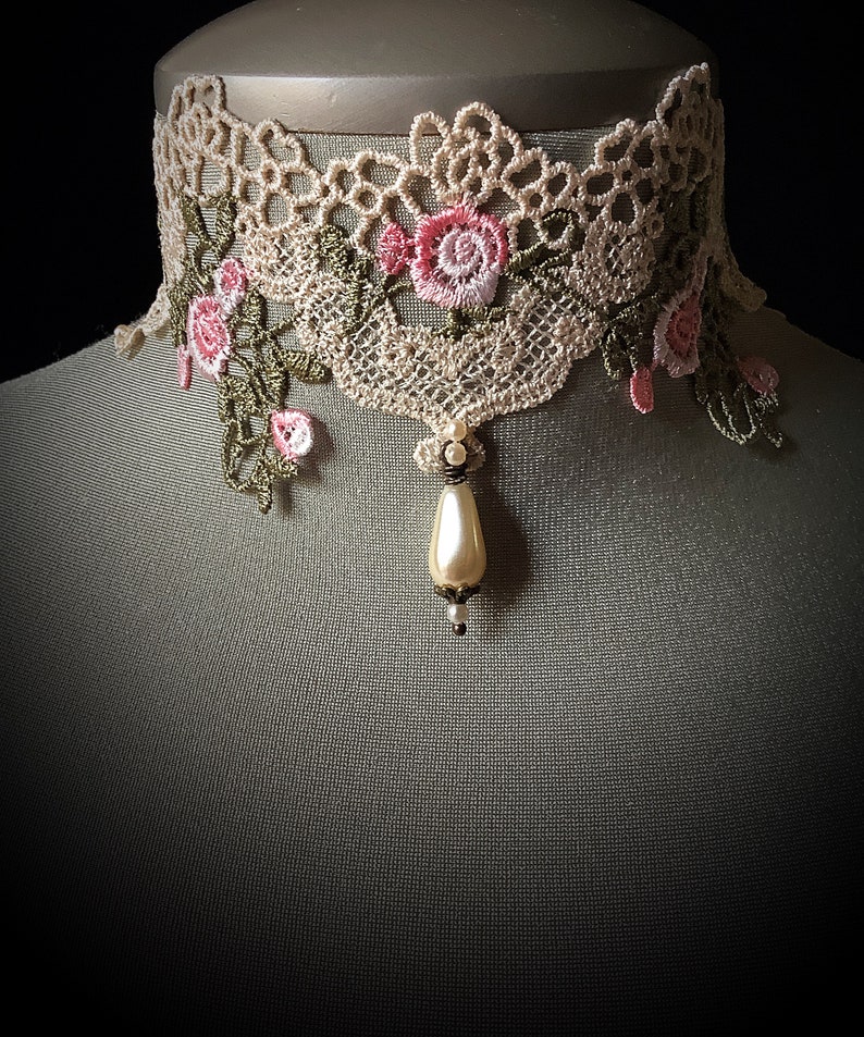 Puede incluir: Un delicado collar de encaje beige con bordado floral rosa y un colgante de perla.