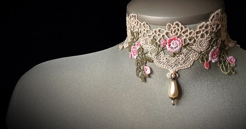 Puede incluir: Un delicado collar de encaje marfil con bordados florales rosas y verdes y un colgante de perla.