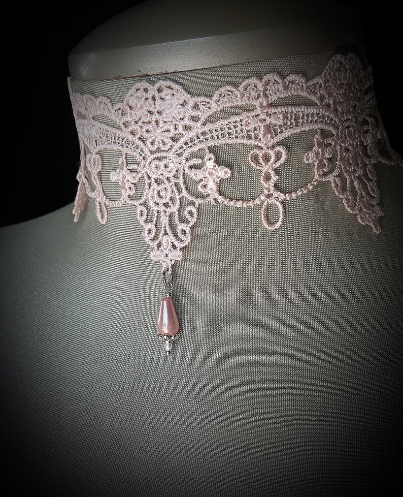 Puede incluir: Un delicado collar gargantilla de encaje rosa claro con intrincados patrones florales y de volutas. Un colgante de cuentas rosa en forma de pera cuelga del centro. El collar se exhibe en un maniqu&iacute;.