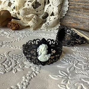 May include: Two black metal hair clips with a cameo design. The cameo features a white porcelain profile of a woman with a floral design.