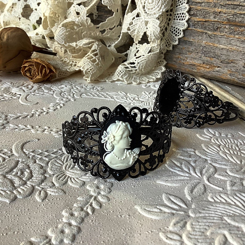 May include: Two black metal hair clips with a cameo design. The cameo features a white porcelain profile of a woman with a floral design.