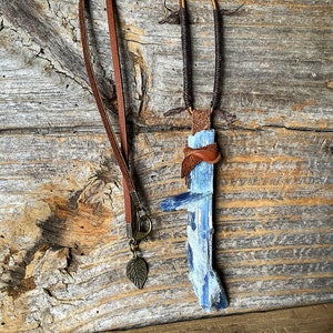 Puede incluir: Un colgante de cristal de cianita azul en un collar de cordón de cuero marrón. El colgante está envuelto en cuero marrón y tiene un cierre de metal.