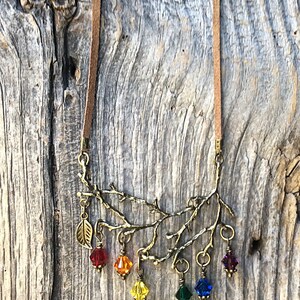 May include: A necklace featuring a brown suede cord and a bronze-colored branch pendant. The pendant has a leaf charm and dangling, colorful crystal beads in red, orange, yellow, green, blue, and purple. The necklace is displayed against a weathered wood background.