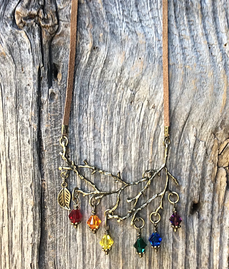 May include: A necklace featuring a brown suede cord and a bronze-colored branch pendant. The pendant has a leaf charm and dangling, colorful crystal beads in red, orange, yellow, green, blue, and purple. The necklace is displayed against a weathered wood background.