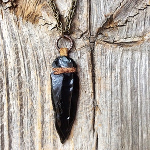 Puede incluir: Un collar con un colgante de punta de flecha de obsidiana negra, con una envoltura de cuero marr&oacute;n y una cadena de bronce.