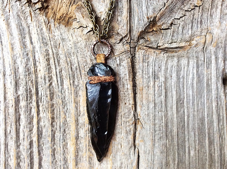 Puede incluir: Un collar con un colgante de punta de flecha de obsidiana negra, con una envoltura de cuero marr&oacute;n y una cadena de bronce.