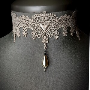 May include: A delicate lace choker with a pearlescent pendant. The choker is made of beige lace and features a delicate floral pattern. The pendant is a teardrop shape and is attached to the choker with a silver chain.
