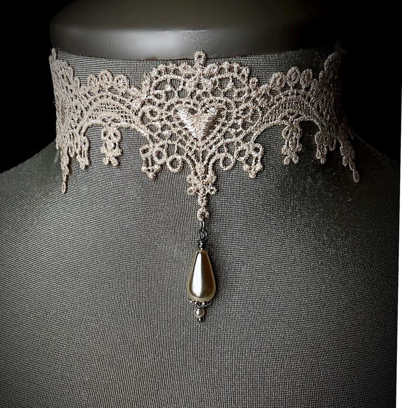 May include: A delicate lace choker with a pearlescent pendant. The choker is made of beige lace and features a delicate floral pattern. The pendant is a teardrop shape and is attached to the choker with a silver chain.