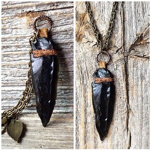 Puede incluir: Un collar con un colgante de punta de flecha de obsidiana negra, con una envoltura de cuero marr&oacute;n y detalles en cobre. El collar est&aacute; en una cadena con un dije de hoja.