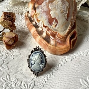 May include: A silver ring with a blue cameo featuring a woman's profile. The ring is on a white surface with a seashell and dried roses.