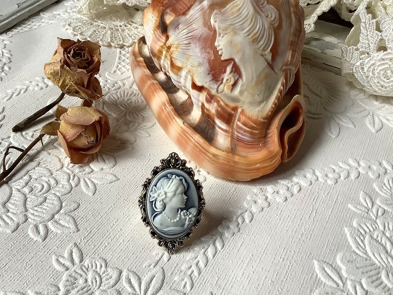 May include: A silver ring with a blue cameo featuring a woman's profile. The ring is on a white surface with a seashell and dried roses.