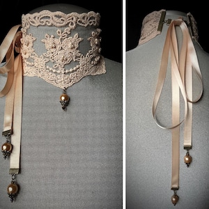 May include: A delicate, peach-colored lace choker necklace with a floral design and pearl accents. The choker features satin ribbons and dangling gold-toned pearl pendants, creating an elegant and vintage aesthetic.