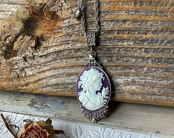 Op de afbeelding: Een zilveren ketting met een cameo-hanger met het profiel van een witte vrouw op een paarse achtergrond. De cameo is omgeven door sierlijke zilveren details.